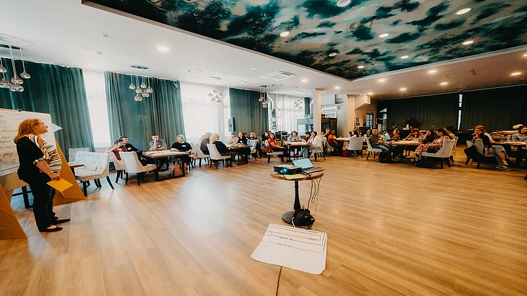 A section of an indoor hall hosting an FES seminar, with a diverse set of participants seated around several large tables and a moderator standing on the left-hand side.