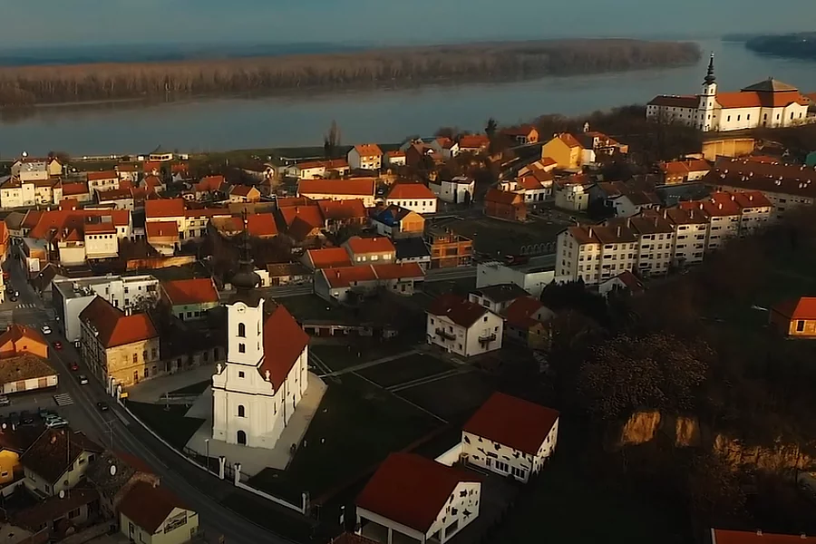 A panoramic photograph of Vukovar shows the city, a sacral (religious) building, and the Danube River, photographed from the air in a wide shot.