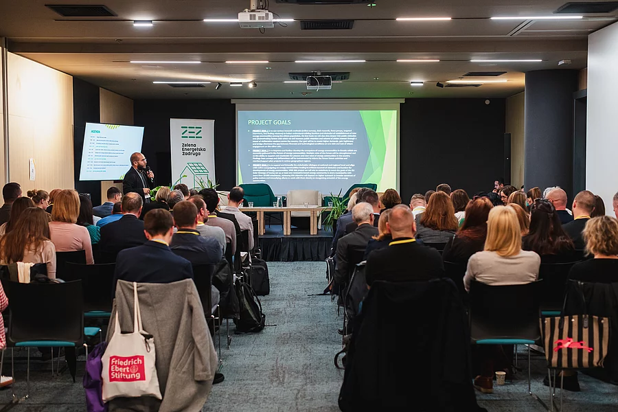 A photograph of a conference hall with the camera facing the stage, showing an empty set of panel chairs, while a speaker in the left corner presents a PowerPoint slide titled “Project Goals” in front of a nearly full auditorium. The FES logo is visible on one of the tote bags in the audience.