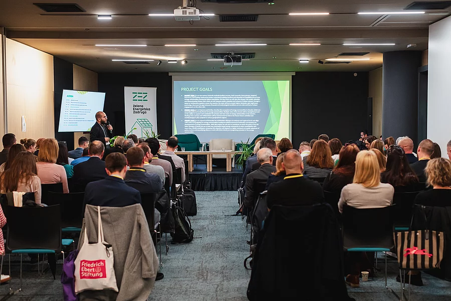 Fotografija konferencijske dvorane snimljena s pogledom prema pozornici, prikazujući prazan niz stolica za paneliste, dok govornik u lijevom kutu prezentira PowerPoint slajd pod nazivom „Ciljevi projekta“ pred gotovo punom dvoranom. Logotip FES-a vidljiv je na jednoj od torbi u publici.