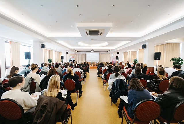 A shot of a large indoor hall with a large audience seated and facing away from the photographer, and a panel with seated panelists and moderators in the distance at the front.