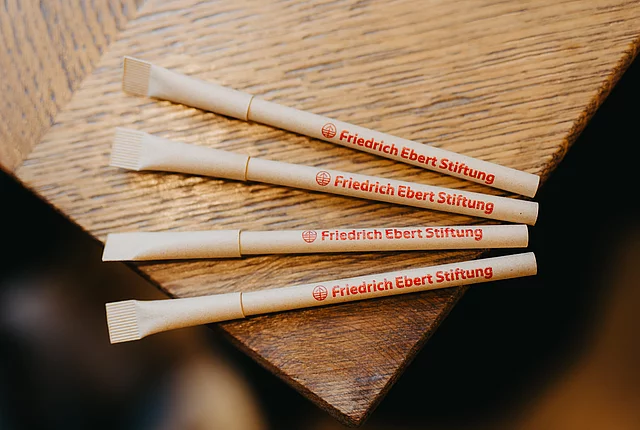 Selection of four pens made from recyclable material with the Friedrich Ebert Stiftung logo on them, placed at the edge of a wooden table.