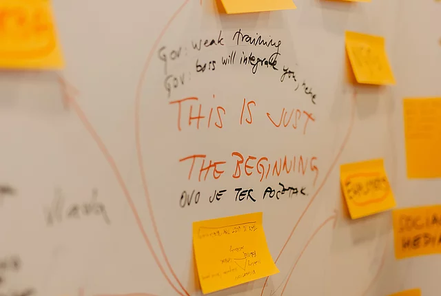A detail shot of a white paper spreadsheet with multiple writings and sticky notes. In the centre, in focus, is a sentence that reads, “This is just the beginning.”