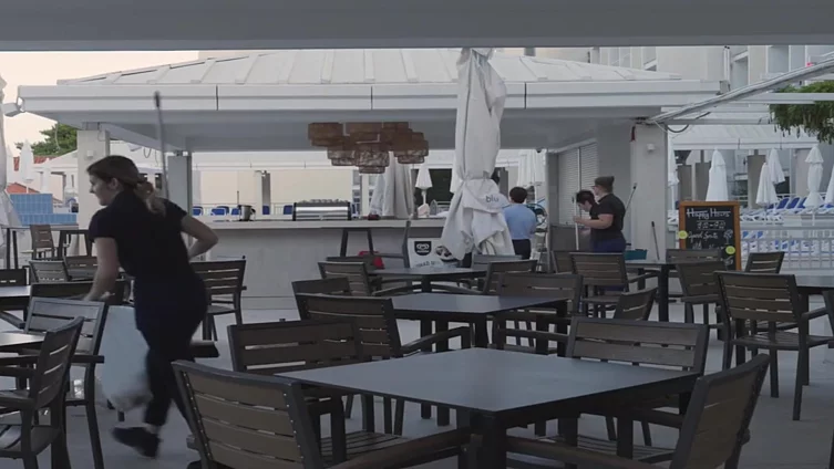 Fotografija ugostiteljskog objekta na obali. Prazni stolovi i stolice, te radnici i radnice pripremaju objekat za otvaranje.