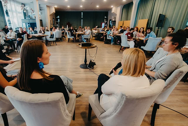 A panel discussion shot from behind the three female panelists, with a large crowd seated around tables at an indoor event visible in the background.