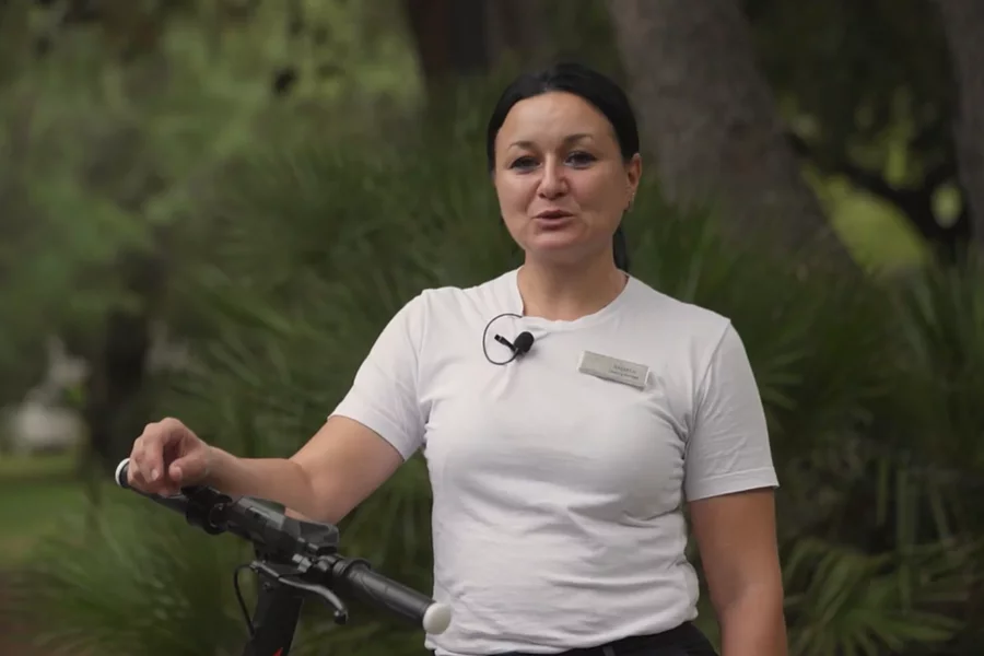 Fotografija žene zaposlenice u turizmu koja gleda u kameru i oslanja se na električni romobil, s cvjetajućom zelenom pozadinom.